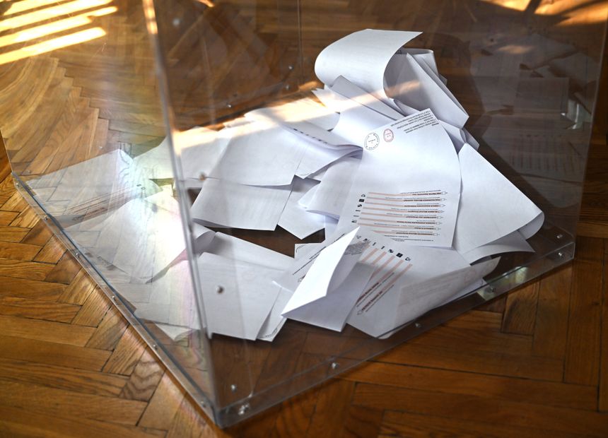 W przezroczystej urnie leżą złożone kartki papieru wyborczego. Drewniana podłoga w tle.