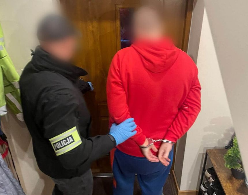 Policjant zatrzymuje osobę w czerwonym ubraniu, przy drzwiach domu. Aresztowany ma założone kajdanki.