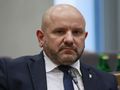 Zdjęcie mężczyzny w garniturze, z krótką brodą, siedzącego w sali obrad.