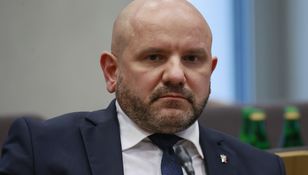 Zdjęcie mężczyzny w garniturze, z krótką brodą, siedzącego w sali obrad.