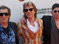 Trzej mężczyźni w okularach przeciwsłonecznych, uśmiechnięci, na tle logo zespołu Rolling Stones.