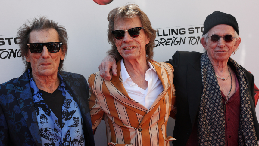 Trzej mężczyźni w okularach przeciwsłonecznych, uśmiechnięci, na tle logo zespołu Rolling Stones.