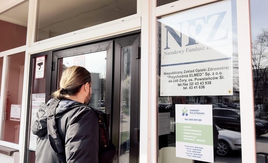 Na zdjęciu mężczyzna stoi przed drzwiami placówki NFZ, spoglądając w kierunku wejścia.