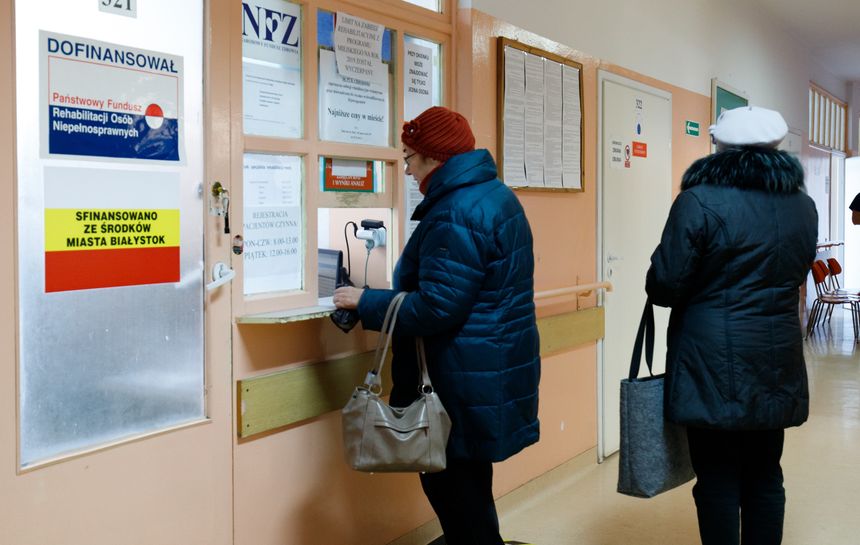 Dwie osoby w kurtkach czekają przy okienku rejestracyjnym w placówce medycznej.