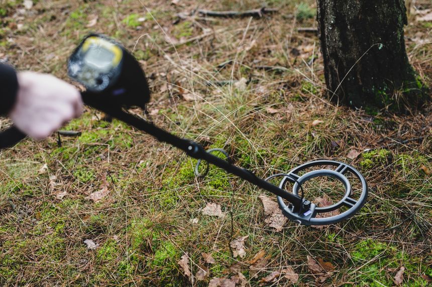 Detektor metali w lesie, trzymany w ręce, na tle leśnego krajobrazu z mchem i igliwiem.