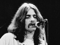 Glenn Frey z Eagles. Mężczyzna z długimi włosami i wąsami śpiewa do mikrofonu na scenie. Czarno-biała fotografia.