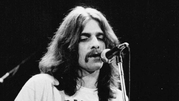 Glenn Frey z Eagles. Mężczyzna z długimi włosami i wąsami śpiewa do mikrofonu na scenie. Czarno-biała fotografia.