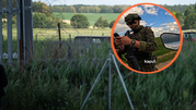 Żołnierz w mundurze trzyma telefon, za nim widok na zielone pola i druty kolczaste.