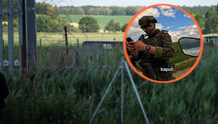 Żołnierz w mundurze trzyma telefon, za nim widok na zielone pola i druty kolczaste.