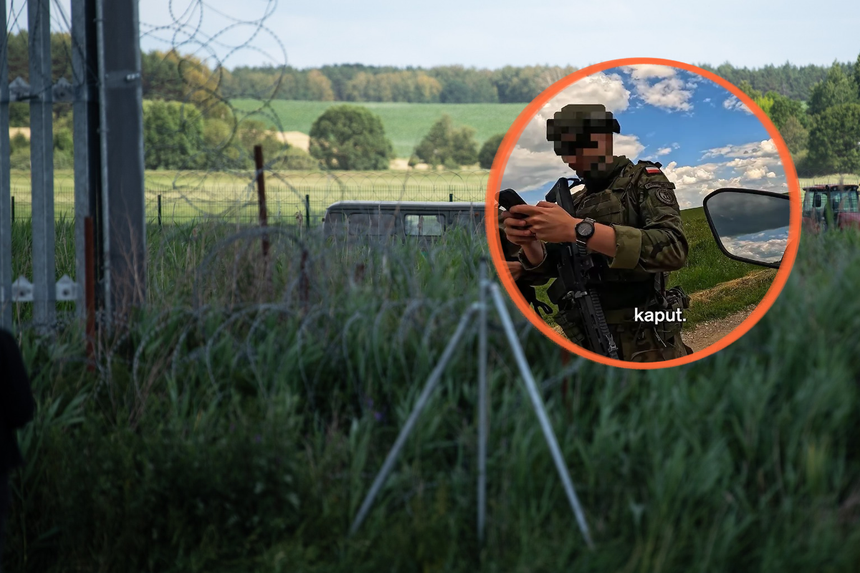 Żołnierz w mundurze trzyma telefon, za nim widok na zielone pola i druty kolczaste.