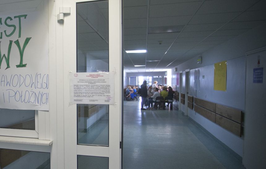 Zdjęcie przedstawia korytarz w placówce medycznej z grupą ludzi w tle. Na ścianie plakat.