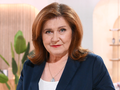 Kobieta o rudych włosach i niebieskich oczach, ubrana w granatowy żakiet, w studio telewizyjnym.