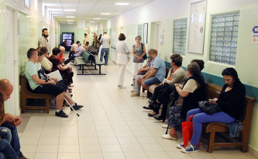 Korytarz w szpitalu z pacjentami czekającymi na wizytę lekarza, personel medyczny w tle.