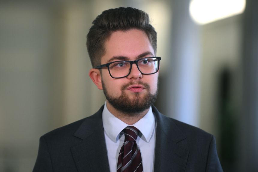 Młody mężczyzna w okularach, ubrany w garnitur, mówi do mikrofonu w jasnym otoczeniu.