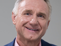 Dan Brown. Mężczyzna w garniturze, uśmiechający się do kamery na tle jasnej ściany.