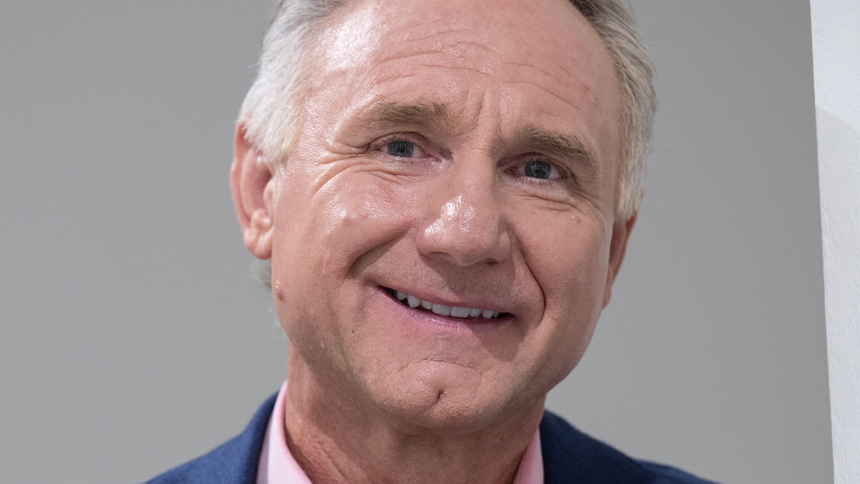 Dan Brown. Mężczyzna w garniturze, uśmiechający się do kamery na tle jasnej ściany.