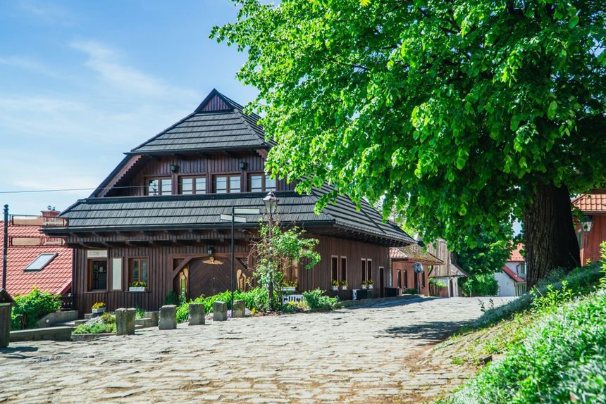 Zdjęcie przedstawia drewniany dom przy kamiennej uliczce, otoczony zielenią drzew.