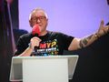 Mężczyzna z okularami i koszulką, trzyma mikrofon, przemawia na scenie.