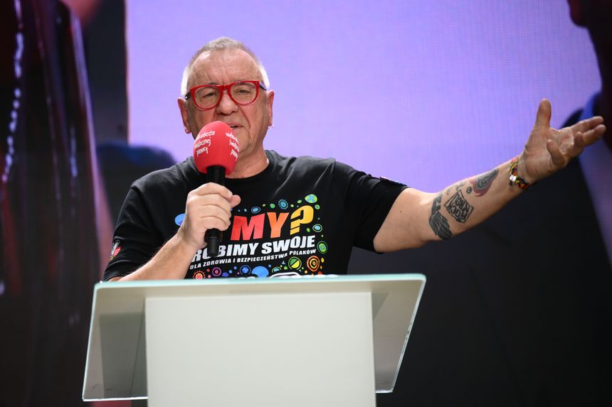 Mężczyzna z okularami i koszulką, trzyma mikrofon, przemawia na scenie.