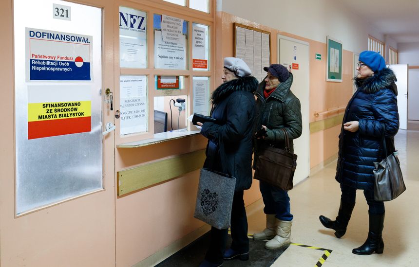 Trzy osoby w zimowych ubraniach stoją przy oknie rejestracyjnym w placówce zdrowotnej.