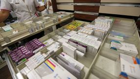Na zdjęciu widać półkę z lekami w aptece, a obok stoi farmaceutka trzymająca preparat.