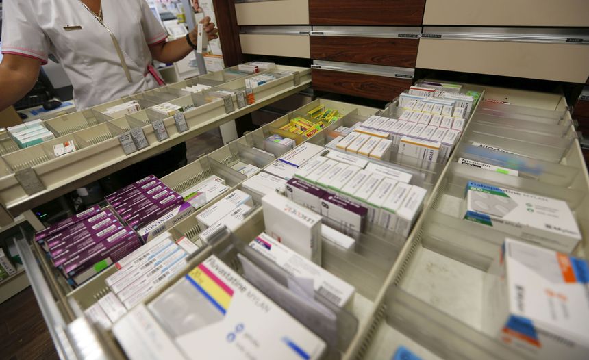 Na zdjęciu widać półkę z lekami w aptece, a obok stoi farmaceutka trzymająca preparat.
