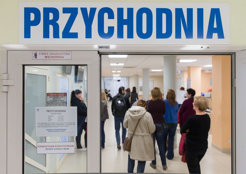 Przychodnia lekarska z osobami w kolejce, widok z drzwi wejściowych. W tle jasne wnętrze.