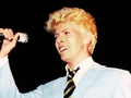 David Bowie. Mężczyzna z jasnymi włosami, w jasnym garniturze, trzyma mikrofon i uśmiecha się.