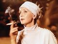Kobieta w białej sukni i turbanie, trzymająca papierosa, z zamyśloną miną. Tło rozmyte.