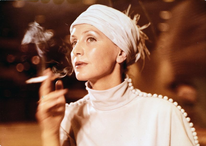 Kobieta w białej sukni i turbanie, trzymająca papierosa, z zamyśloną miną. Tło rozmyte.