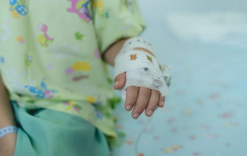 Dziecięca dłoń z opatrunkiem, w szpitalnym ubraniu, z kroplówką na łóżku pediatrycznym.