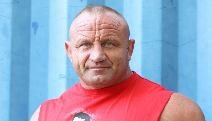 Mariusz Pudzianowski w czerwonym podkoszulku, stojący przed niebieskim tłem, uśmiecha się.