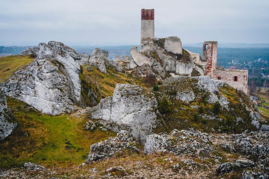 Ruiny na wzgórzu otoczone skałami i zielenią, w tle widok na dolinę, pochmurne niebo.