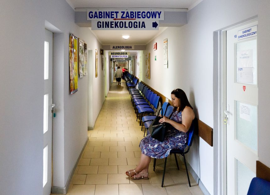 Korytarz w przychodni z niebieskimi krzesłami i kobietą czekającą na wizytę ginekologiczną.