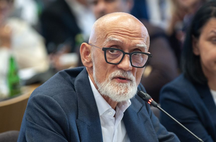 Mężczyzna z siwą brodą i okularami, siedzący przy stole konferencyjnym.