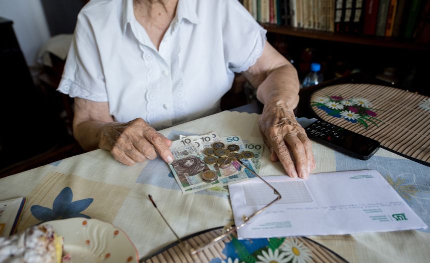 Na stole leżą banknoty i monety, ręce starszej kobiety trzymają pieniądze i okulary.