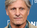 Viggo Mortensen. Mężczyzna z jasnymi włosami, w szarej kurtce, patrzy prosto w obiektyw.