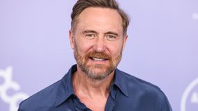 David Guetta w niebieskiej koszuli, z brodą i krótkimi włosami, uśmiecha się na tle purpurowym.