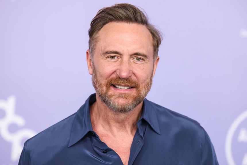 David Guetta w niebieskiej koszuli, z brodą i krótkimi włosami, uśmiecha się na tle purpurowym.