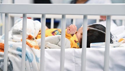 Dziecko w szpitalnym łóżku, owinięte w koc, z widoczną ręką i monitorującym sprzętem.