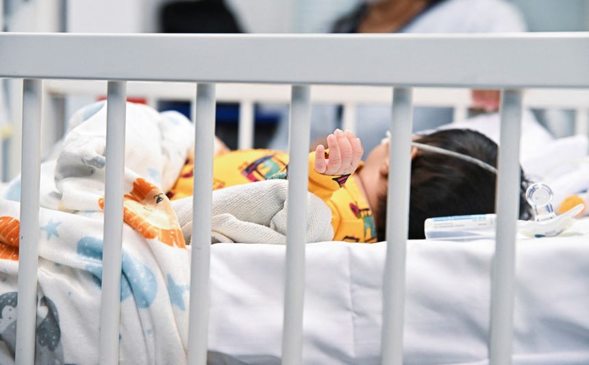 Dziecko w szpitalnym łóżku, owinięte w koc, z widoczną ręką i monitorującym sprzętem.