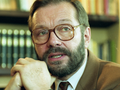 Mężczyzna z brodą i okularami, rozmawia, ręce złożone pod brodą, w tle półki z książkami.
