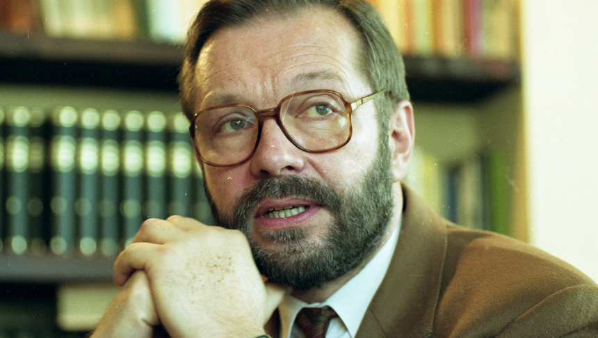Mężczyzna z brodą i okularami, rozmawia, ręce złożone pod brodą, w tle półki z książkami.