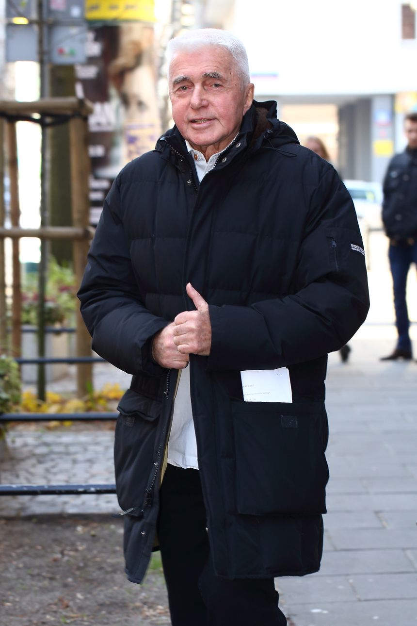 Mężczyzna w czarnej kurtce z białą koszulą, spacerujący po mieście w słoneczny dzień.