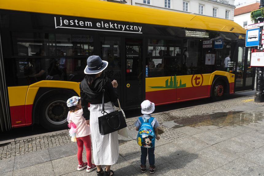 Na przystanku autobusowym stoją kobieta i dzieci, obok elektryczny autobus z napisem „jestem elektryczny”.