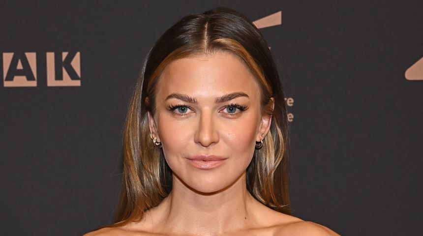 Anna Lewandowska z długimi włosami, uśmiechająca się na tle ciemnej ścianki z logo wydarzenia.