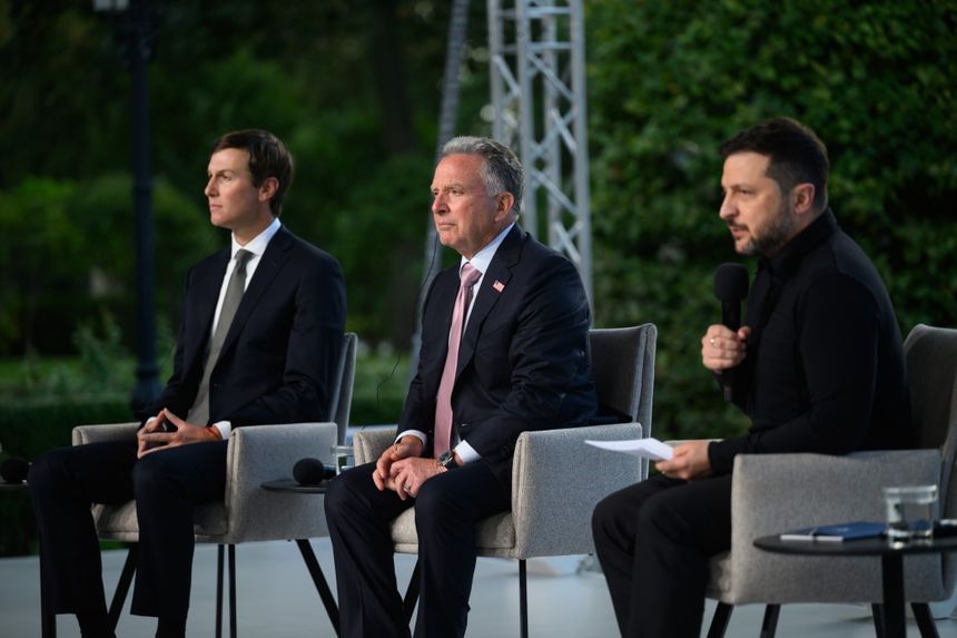 Trzech mężczyzn siedzi na krzesłach, rozmawiając przy stole na tle zieleni.