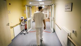 Mężczyzna w białym fartuchu idzie korytarzem szpitalnym. Ściany żółte, sprzęt medyczny w tle.