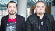 Para mężczyzn w eleganckich ubraniach, stojących blisko siebie w jasnym wnętrzu.