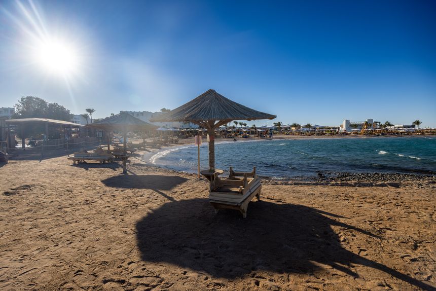Plaża z parasolami, leżakami i falującą wodą, w tle słońce i błękitne niebo.